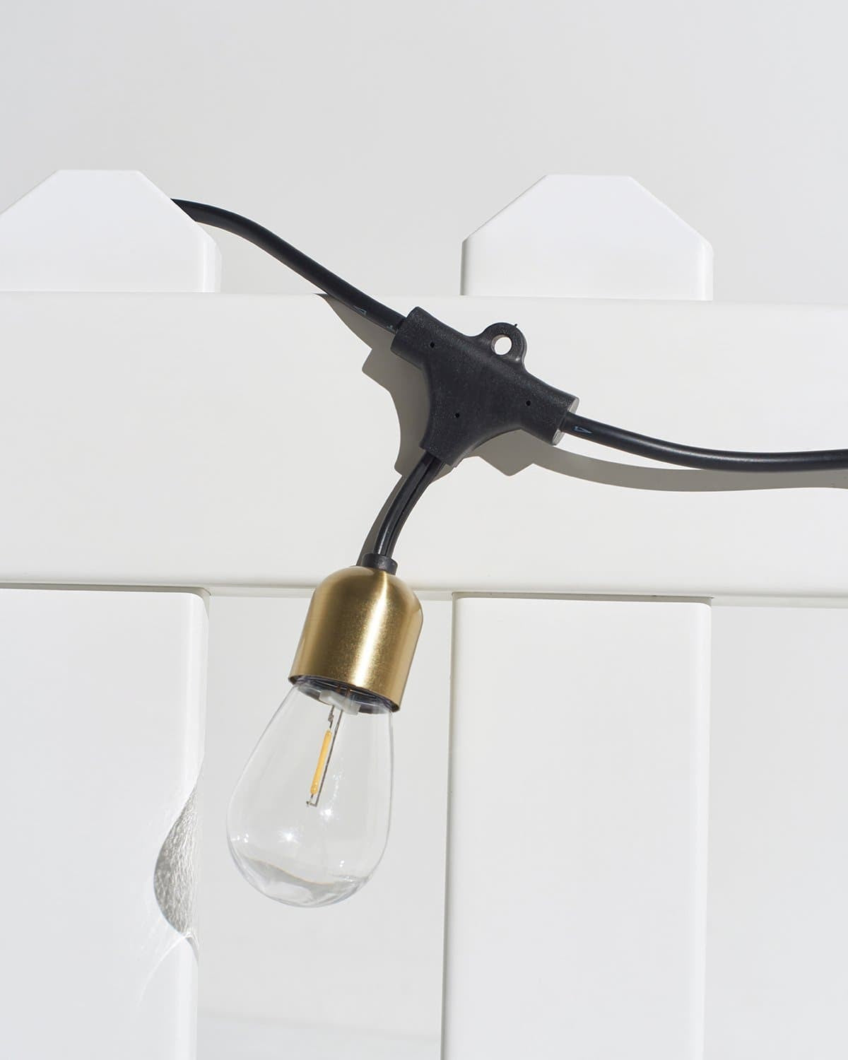 Brightech Glow Solar Hanging string light with a brass-colored socket hanging over a white fence