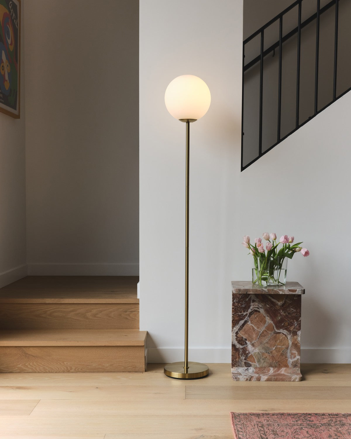 Lit Brightech Luna floor lamp with a frosted glass globe on a brass stem beside a staircase