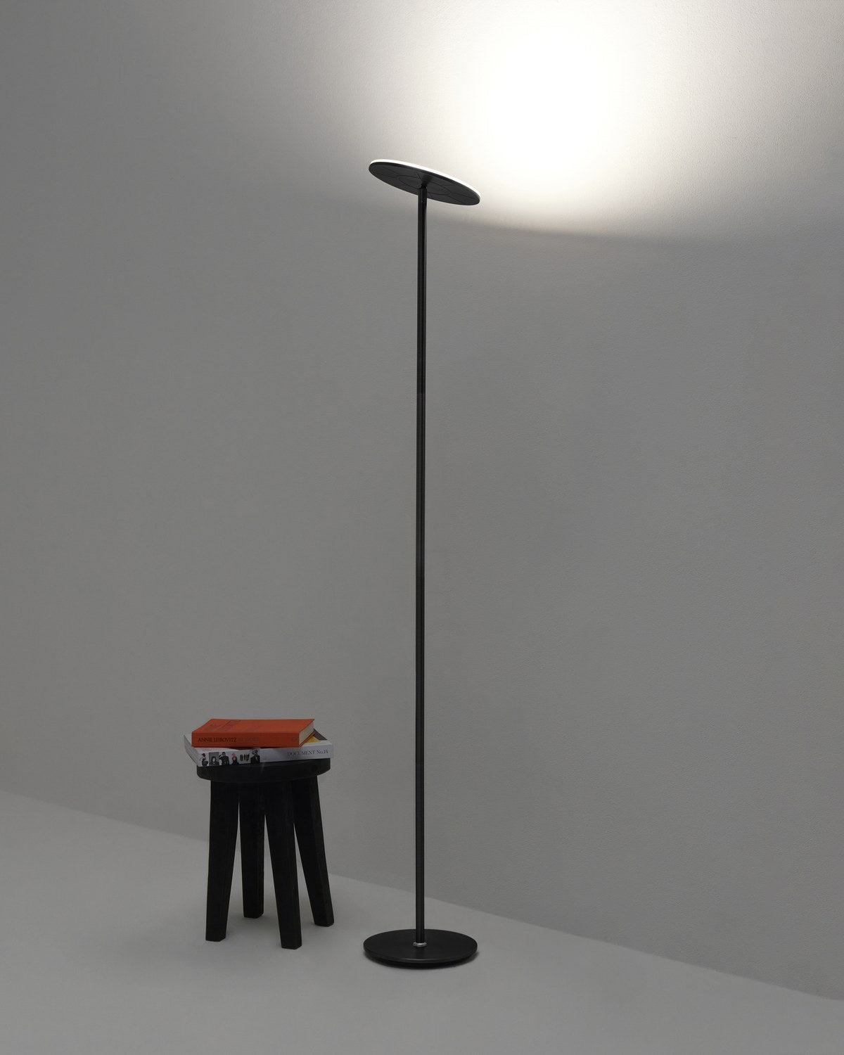 Black Brightech Sky LED torchiere floor lamp washing light up the wall of a dark grey room next to a black stool