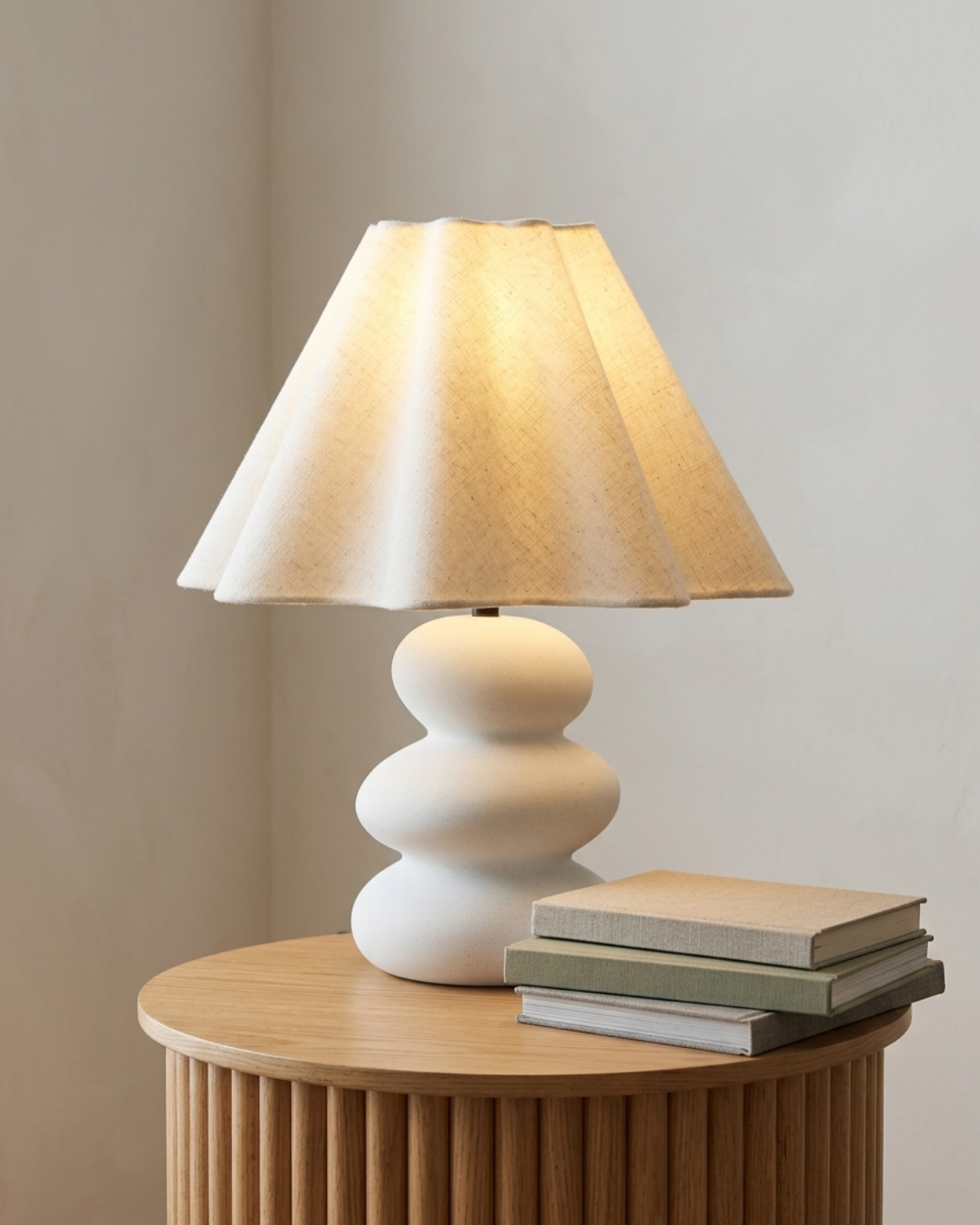A table lamp with a pleated beige fabric shade and a sculptural stacked-white ceramic base, illuminated and styled on a round wooden side table next to a stack of books against a neutral beige wall.