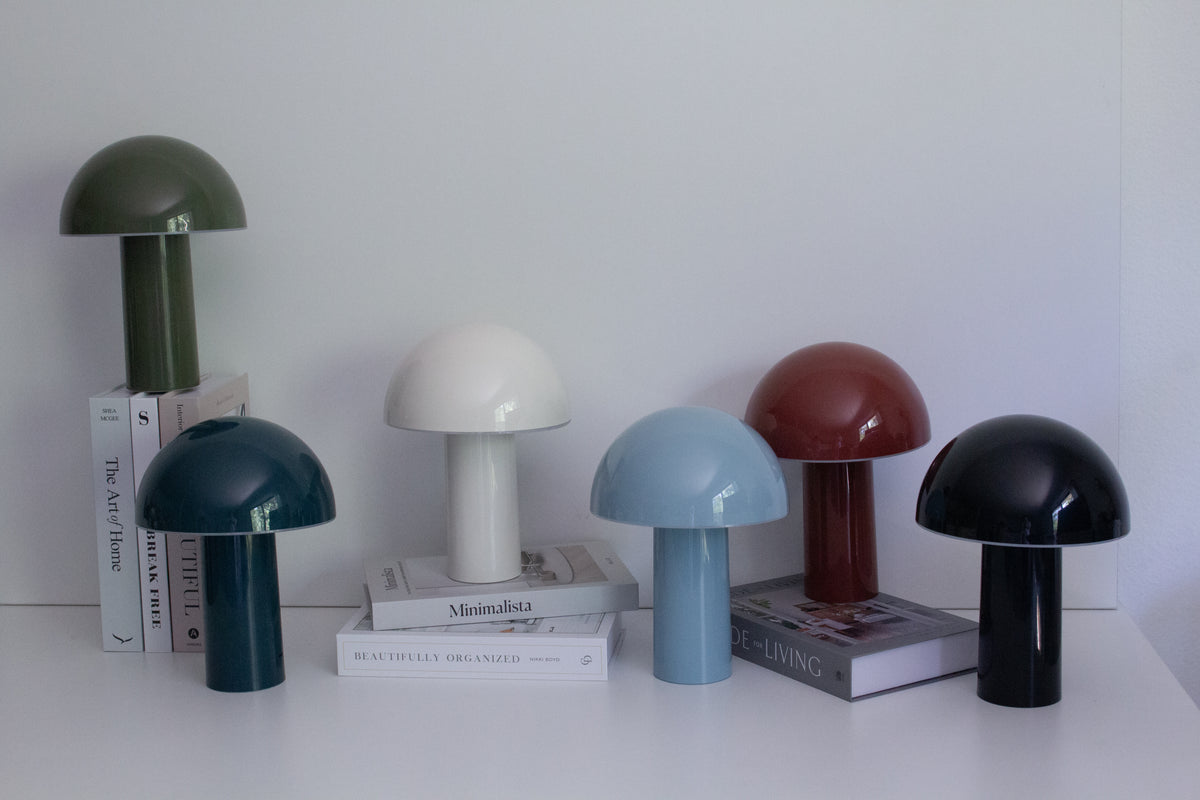 Six Brightech Celia portable lamps in green, teal, cream, light blue, red and black on a white shelf with books