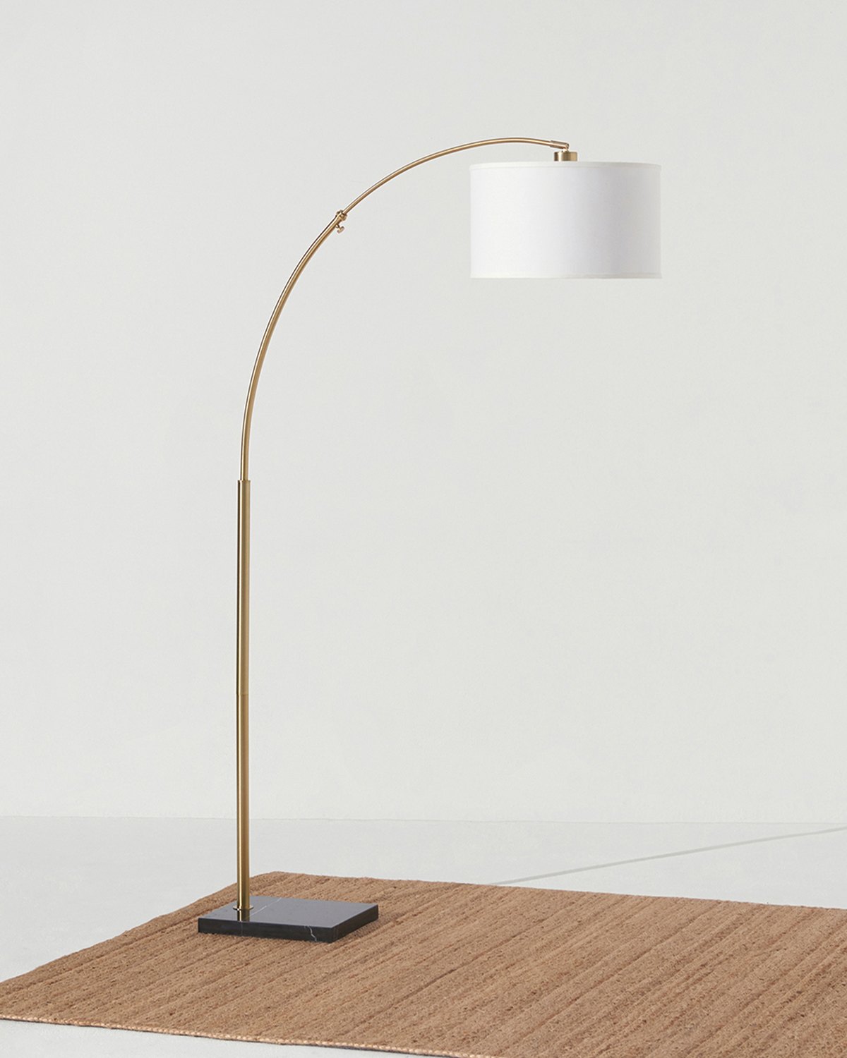 Brass Brightech Logan arc floor lamp with a white drum shade on a black marble base and a jute rug