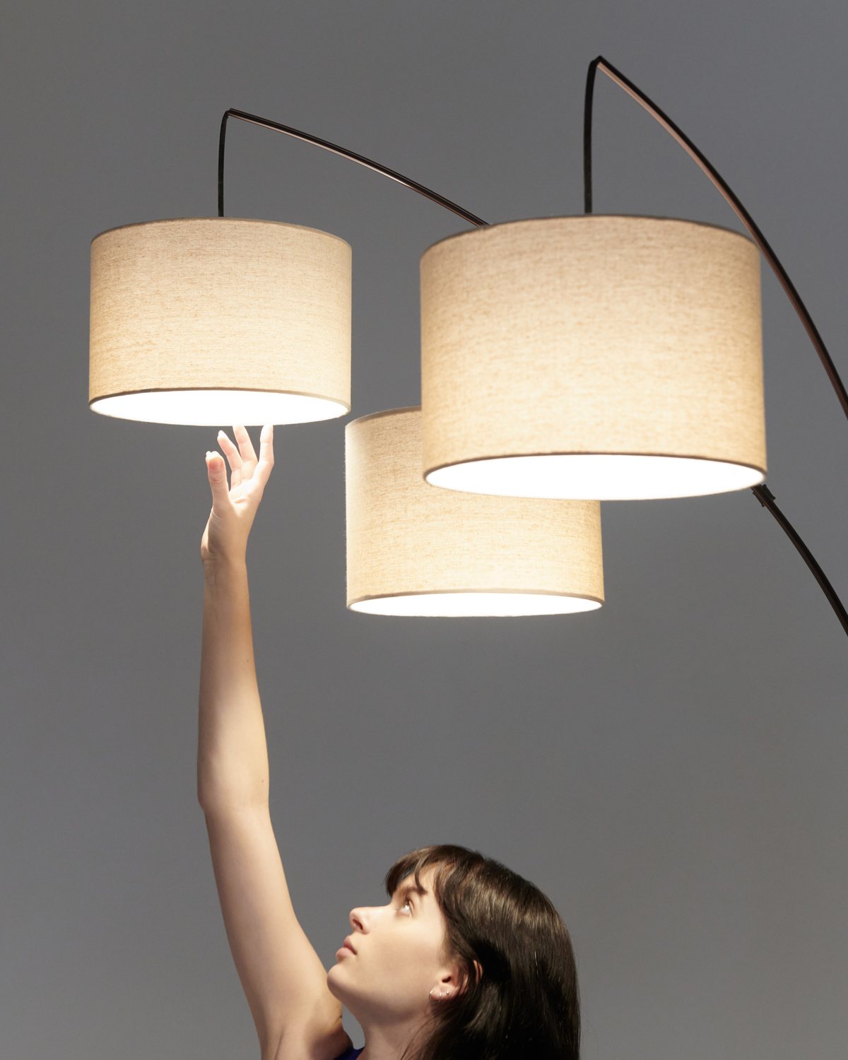 Woman reaching up to the three linen drum shades of the Brightech Trilage floor lamp