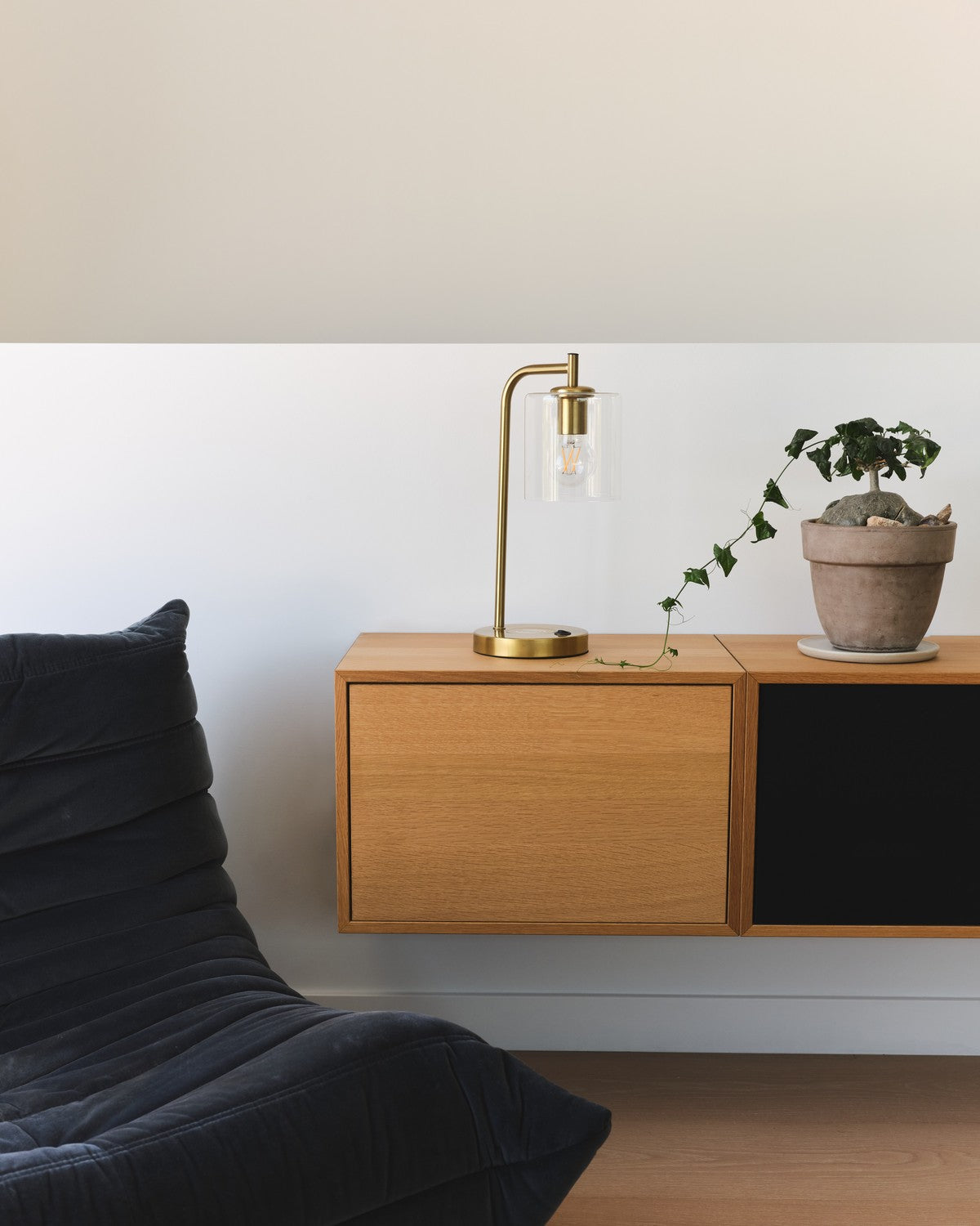 Lit brass Brightech Elizabeth table lamp with a clear glass shade on a floating wood desk beside a plant