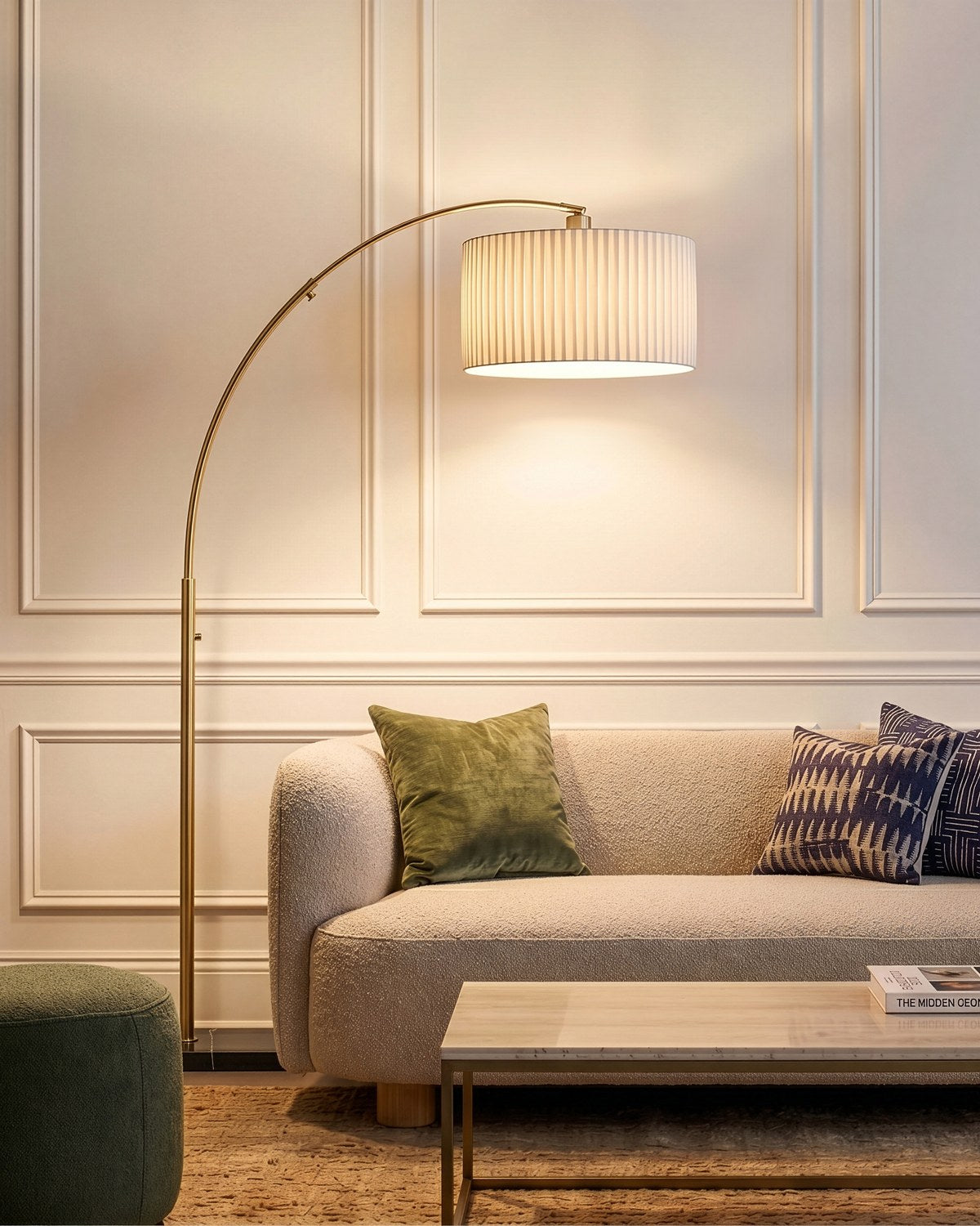 Brightech Logan arc floor lamp in brass with a lit pleated drum shade arching over a cream sofa in a paneled living room