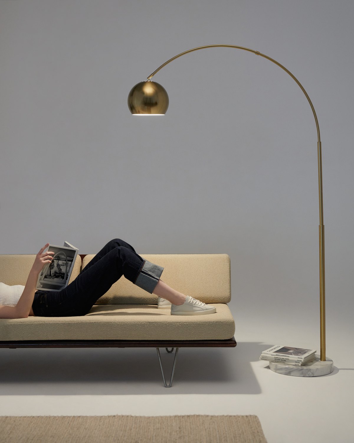 Brass Brightech Olivia arc floor lamp hanging its globe shade over a woman reading on a sofa