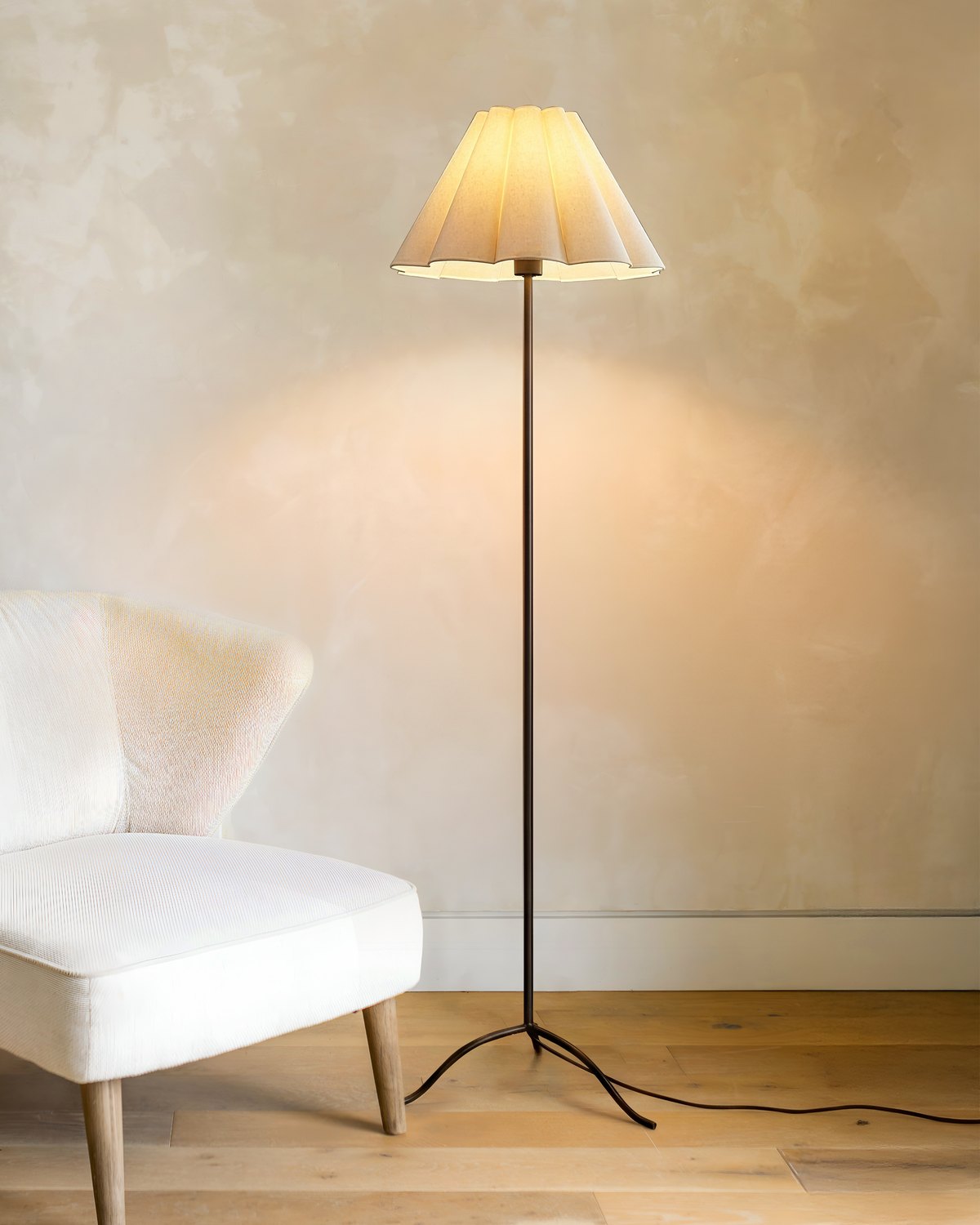 Lit Brightech Lea floor lamp with a fluted empire shade on a slim black tripod stem beside a white armchair