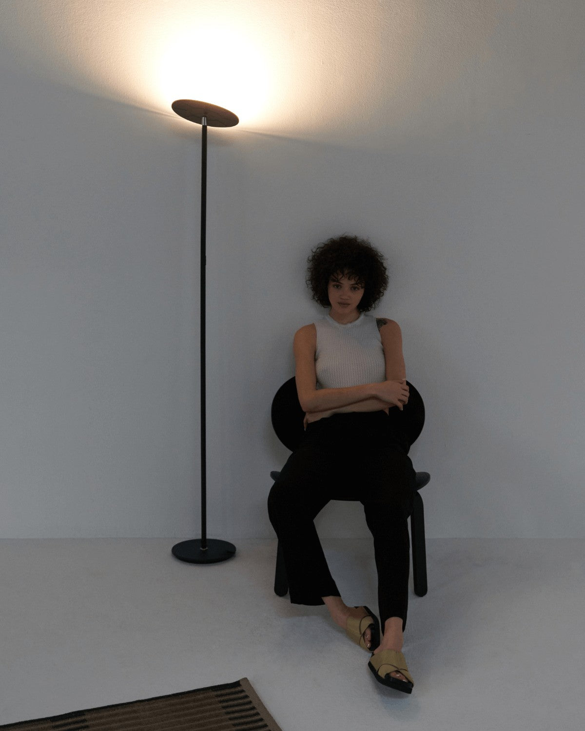 Black Brightech Sky torchiere floor lamp washing a white wall with light above a seated woman
